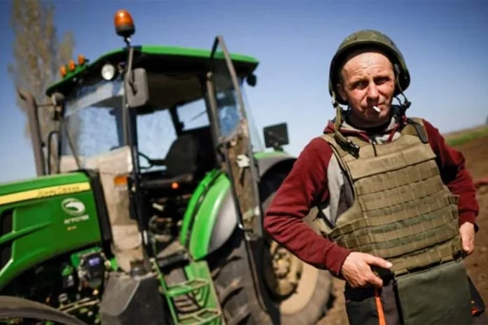 Аграрій у бронежилеті та касці стоїть біля трактора John Deere на полі.