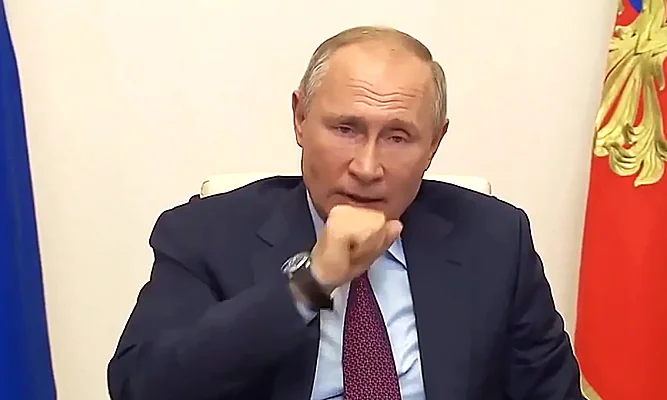 Володимир Путін кашляє, прикриваючи рот кулаком, на тлі прапорів Росії