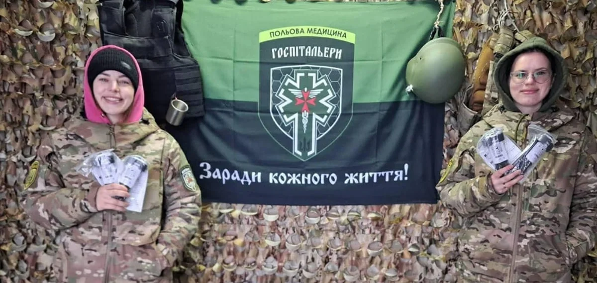 Дві госпітальерки тримають пачки грошей біля прапора батальйону «Госпітальєри» на передовій.