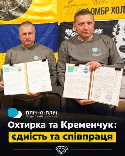 Двоє чоловіків у сірих світшотах тримають підписані документи на тлі жовто-синіх банерів
