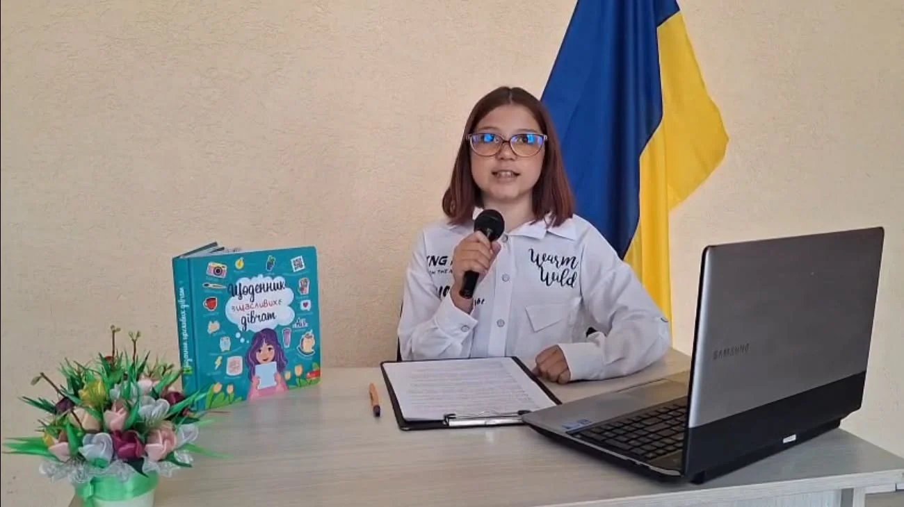 Дівчина з мікрофоном за столом біля книги, ноутбука і прапора України