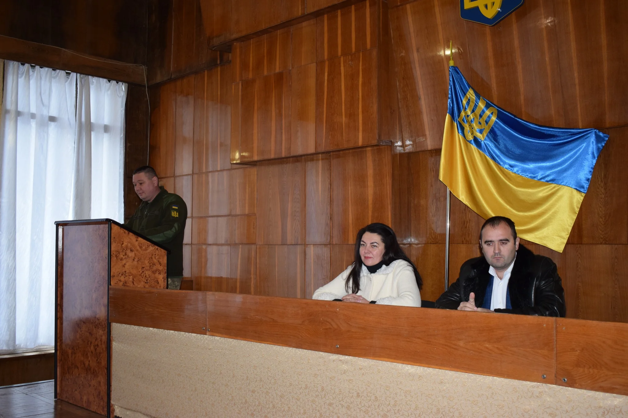 Військовий за трибуною, двоє сидять у залі з українським прапором