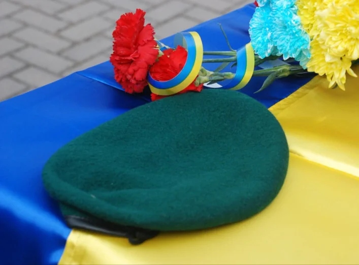 Зелений берет на українському прапорі поруч із червоними гвоздиками та квітами