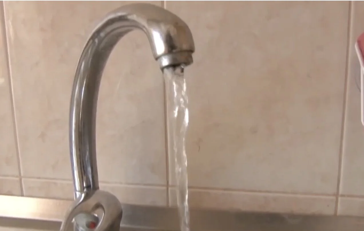 Хромований кухонний кран з якого тече вода на тлі бежевої плитки