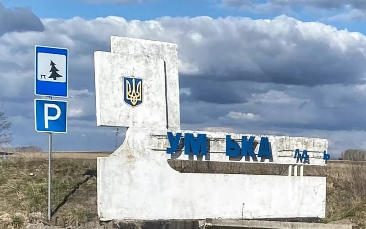 Бетонна стела з гербом України та синіми літерами біля дорожніх знаків на тлі поля